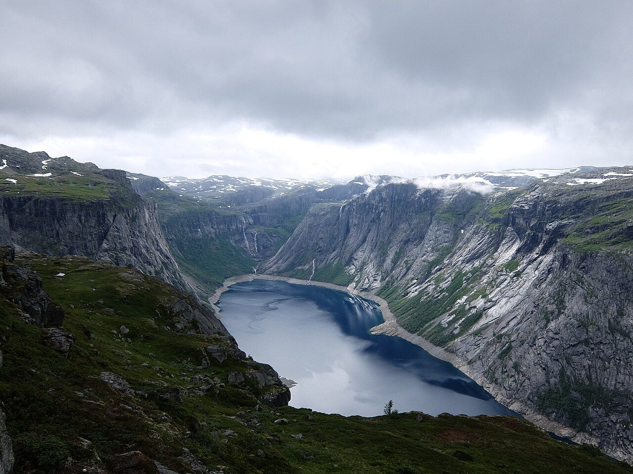 A long blue lake far below steep mountainsides, seen from the plateau at Trolltunga